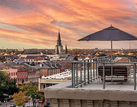 A rooftop deck with a table, umbrella, and chairs overlooks a colorful cityscape at sunset, featuring a church spire and cozy brick buildings.