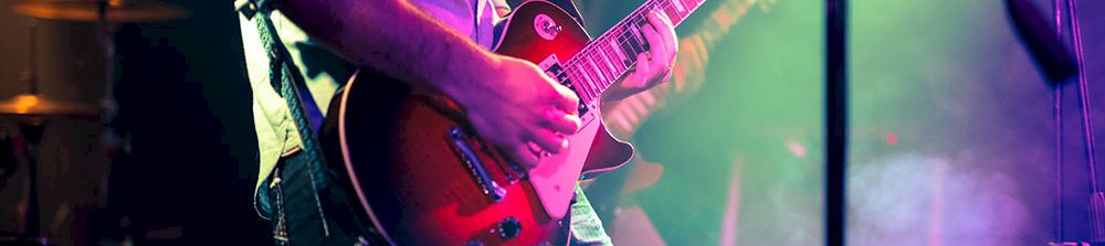 A guitarist on stage plays a red electric guitar under colorful concert lights, with fog and a microphone stand nearby.