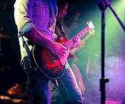 A guitarist on stage plays a red electric guitar under colorful concert lights, with fog and a microphone stand nearby.