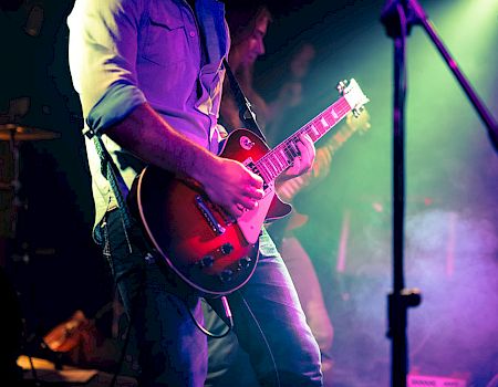 A guitarist on stage plays a red electric guitar under colorful concert lights, with fog and a microphone stand nearby.