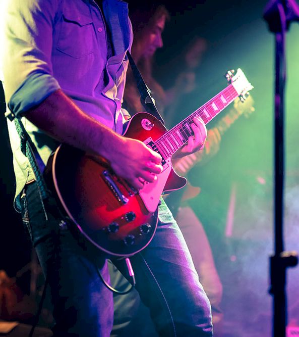 A guitarist on stage plays a red electric guitar under colorful concert lights, with fog and a microphone stand nearby.