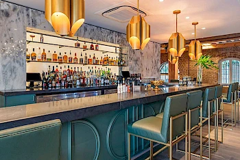 A stylish bar with teal stools, a long marble counter, gold pendant lights, and a well-stocked back bar in a chic, airy space.