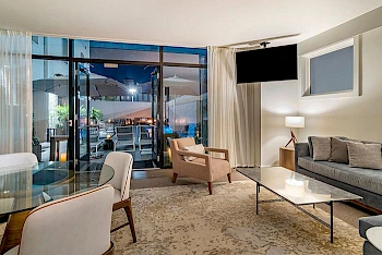 A modern hotel living area with a glass door balcony, sofa, chairs, coffee table, TV, and a warm lamp, overlooking an outdoor seating area at night.