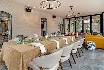 A long dining table set for a meeting or event in a bright, modern restaurant with beige tablecloths, white chairs, greenery, and stylish lighting.