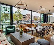 A bright, modern cafe with large windows, cozy couches, wooden tables, pendant lights, and greenery. People and city street visible outside; inviting lounge area.