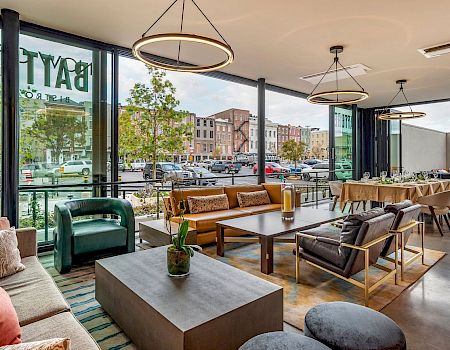 A bright, modern cafe with large windows, cozy couches, wooden tables, pendant lights, and greenery. People and city street visible outside; inviting lounge area.