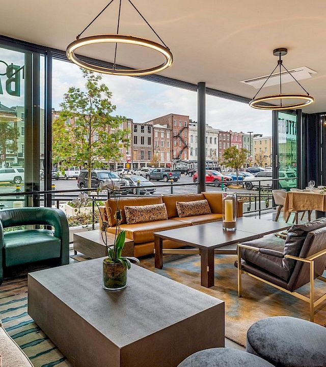 A bright, modern cafe with large windows, cozy couches, wooden tables, pendant lights, and greenery. People and city street visible outside; inviting lounge area.