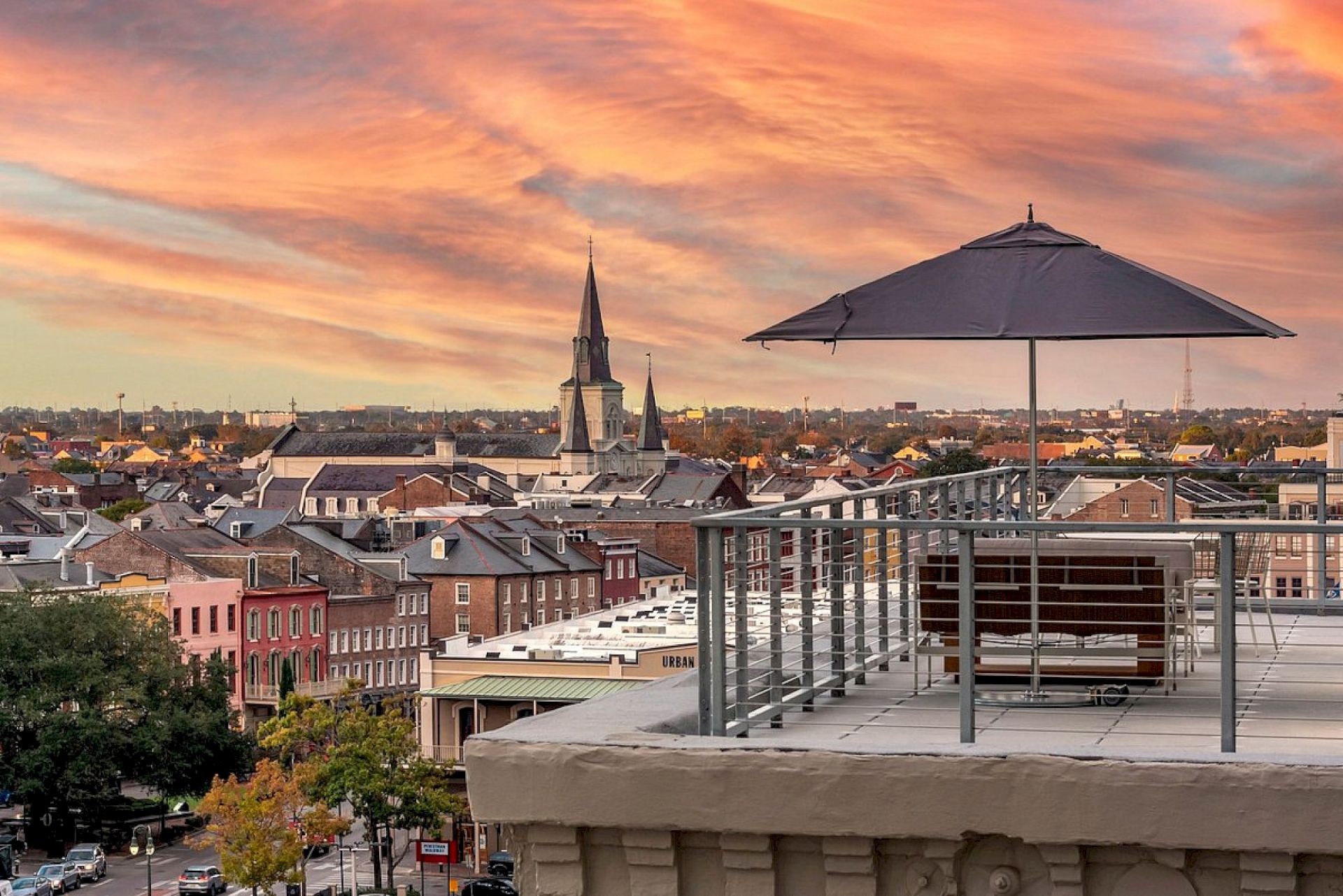 A rooftop terrace with a table, umbrella, and chair overlooking a colorful townscape at sunset.