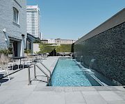 A rooftop pool area at a boutique hotel with clear water, modern lounge chairs, and a gray brick wall on the right; urban buildings loom in the background.