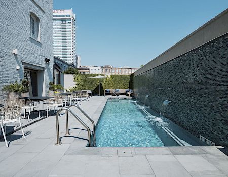 A rooftop pool area at a boutique hotel with clear water, modern lounge chairs, and a gray brick wall on the right; urban buildings loom in the background.