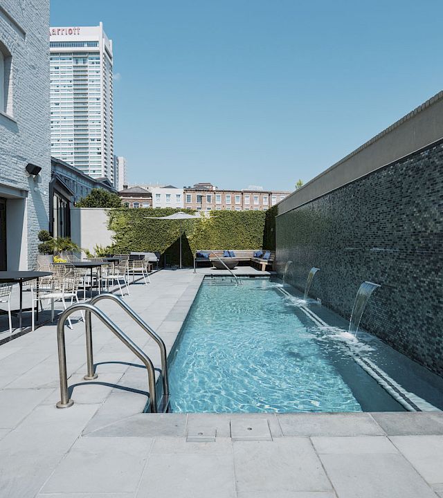 A rooftop pool area at a boutique hotel with clear water, modern lounge chairs, and a gray brick wall on the right; urban buildings loom in the background.
