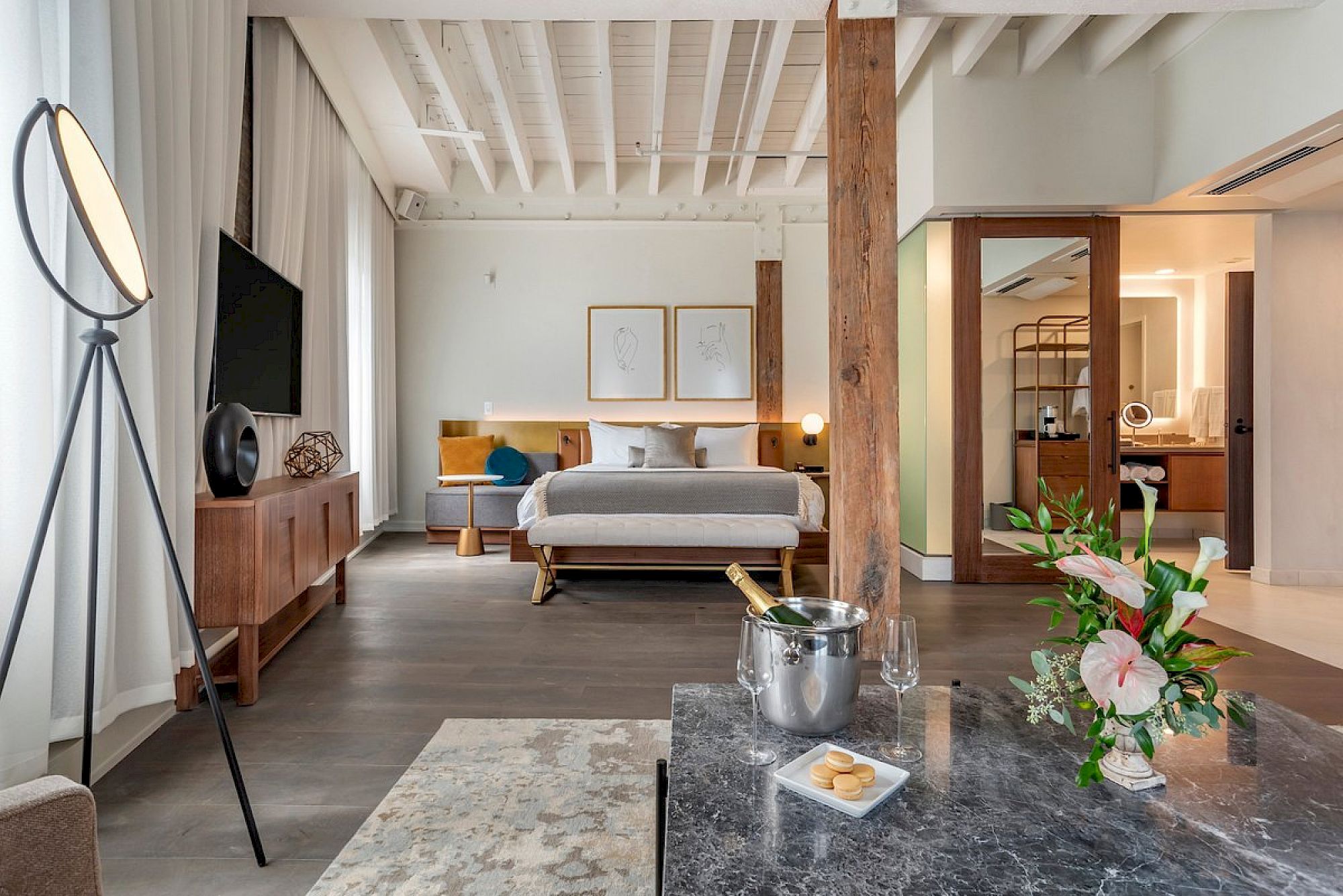 A stylish hotel suite with a king bed, modern living area, warm wood beams, a marble coffee table, flowers, and a cozy, spacious layout.