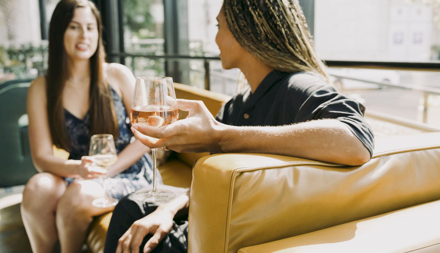Two friends are relaxing on a tan sofa, holding wine glasses and chatting in a bright, modern lounge.