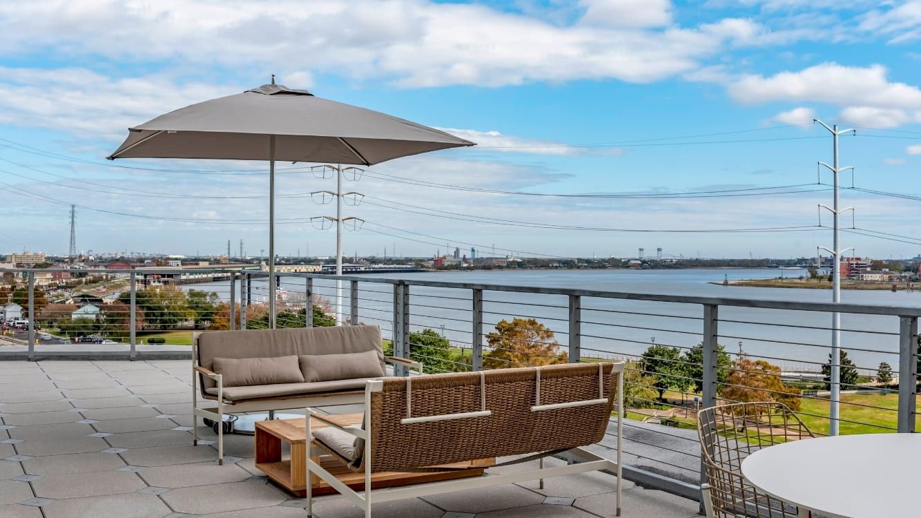 A modern rooftop deck with a beige outdoor sofa and matching chair under a large umbrella, overlooking water and a clear blue sky.