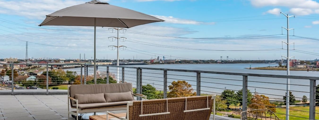 A modern rooftop deck with a beige outdoor sofa and matching chair under a large umbrella, overlooking water and a clear blue sky.