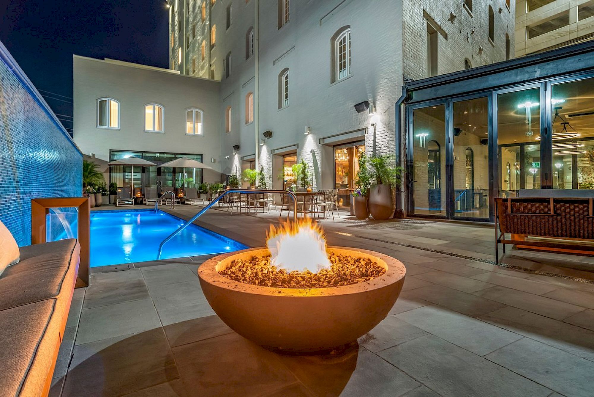 A modern hotel courtyard at night with a lit fire pit, pool, lounge seating, and glass-walled dining area surrounding a stylish exterior space.