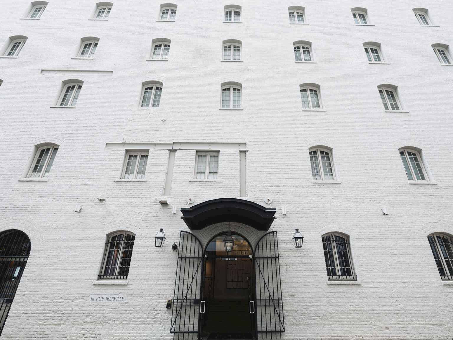 A tall white brick building with many small windows and a central arched door, barred windows, and a secure entrance.