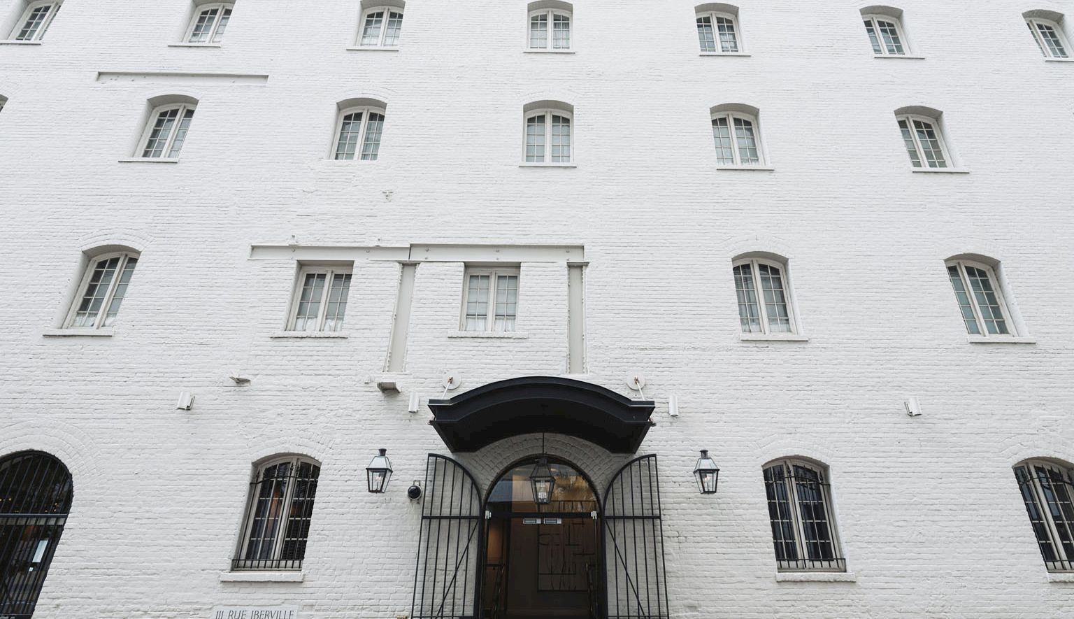 A tall white brick building with many small windows and a central arched door, barred windows, and a secure entrance.