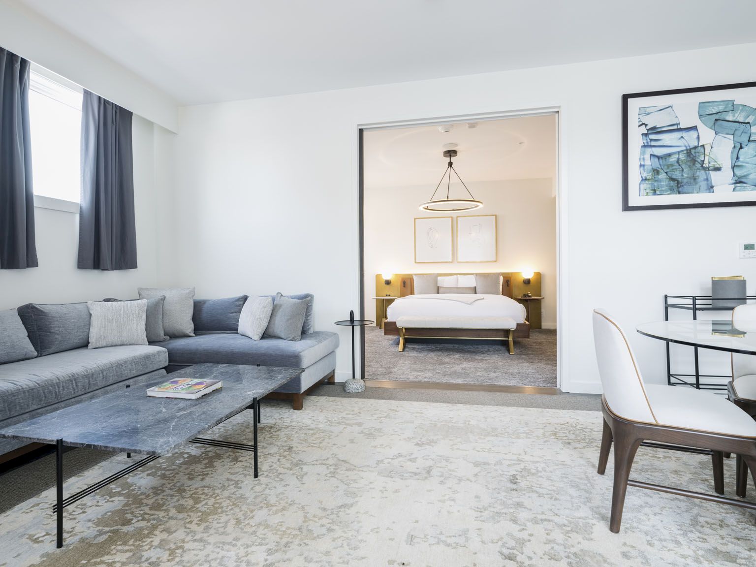 A modern hotel suite with a gray sectional sofa, coffee table, and a rug; through the doorway, a bedroom with a bed, bedside lamps, and a framed art wall.