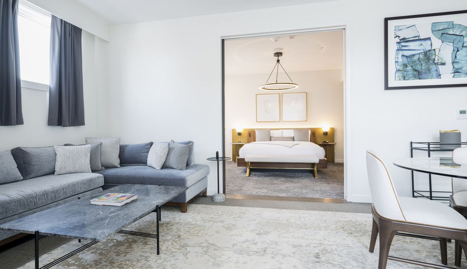 A modern hotel suite with a gray sectional sofa, coffee table, and a rug; through the doorway, a bedroom with a bed, bedside lamps, and a framed art wall.