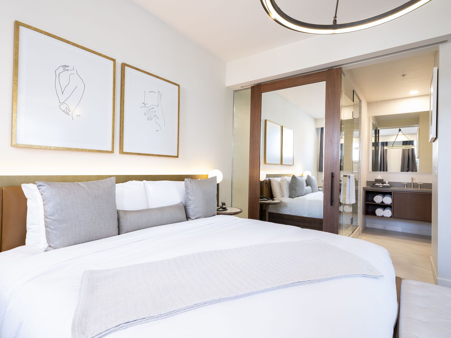 A bright hotel room with a large bed, grey and white bedding, framed minimalist art, and a doorway leading to a glossy, modern bathroom.