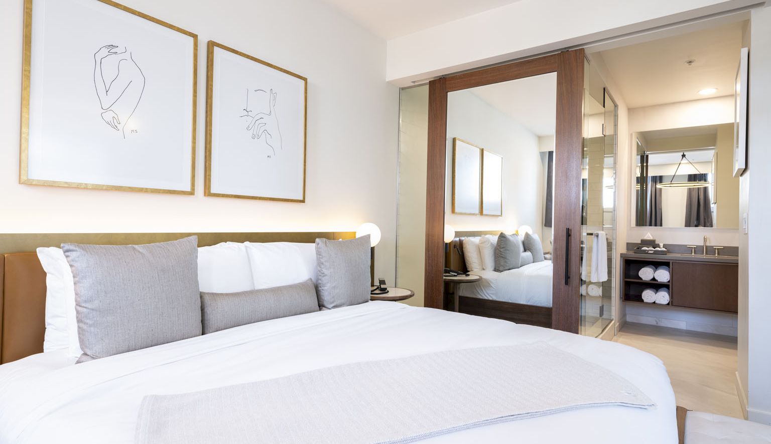A bright hotel room with a large bed, grey and white bedding, framed minimalist art, and a doorway leading to a glossy, modern bathroom.
