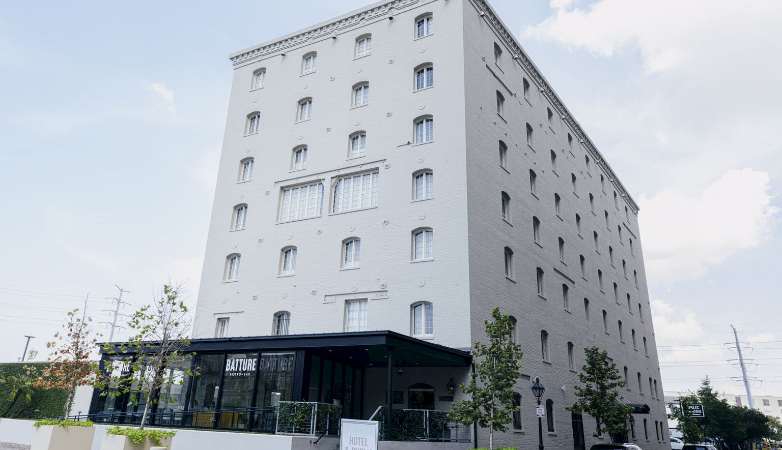 A modern, light-gray, multi-story building with many small windows and a glass-ground-floor entrance; street view with parked cars.