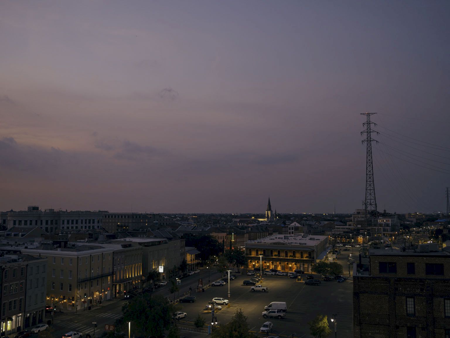 A dusky cityscape with low buildings, a few streetlights, and a tall transmission tower against a dim, purple-tinted sky at dusk.