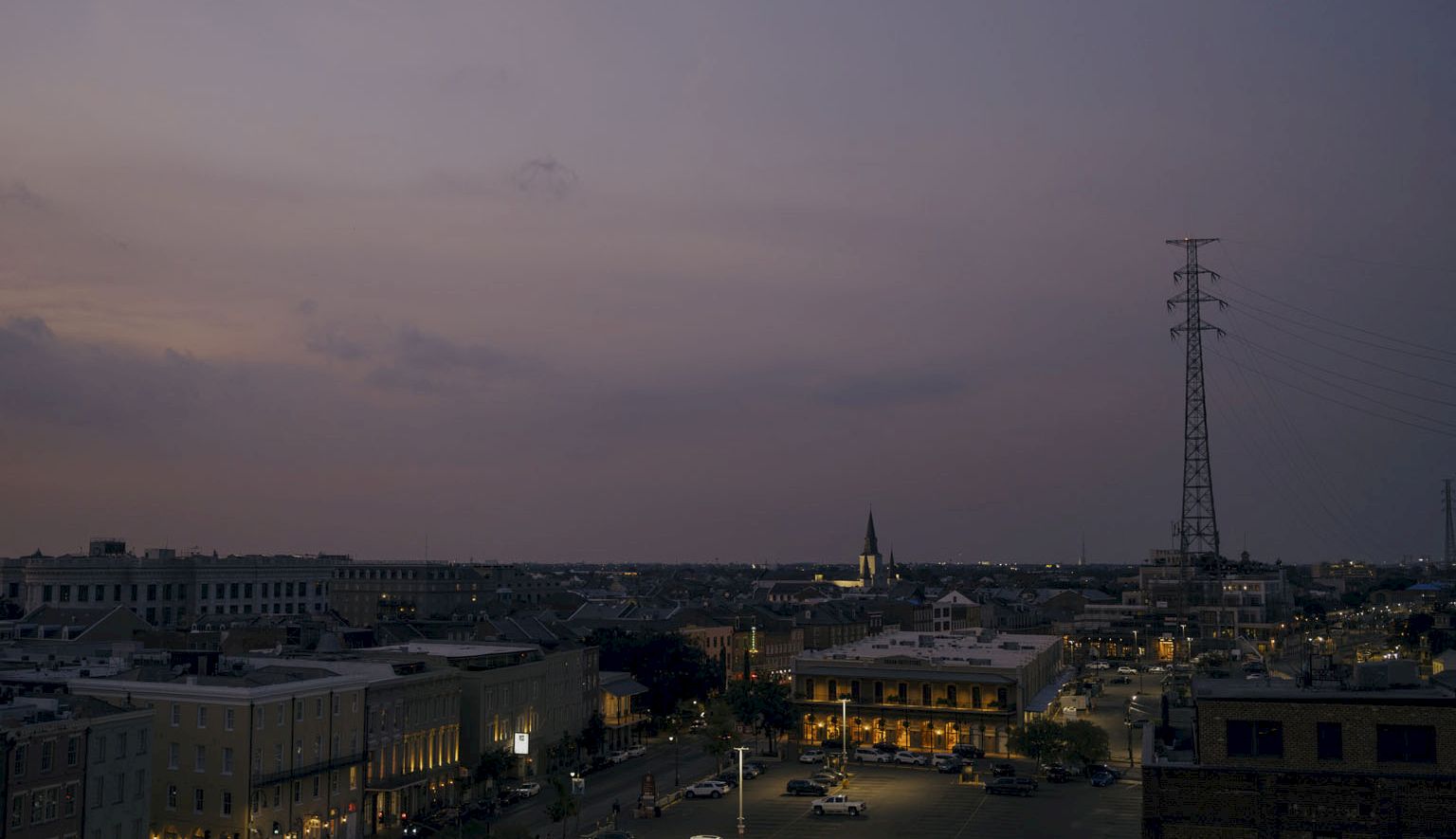 A dusky cityscape with low buildings, a few streetlights, and a tall transmission tower against a dim, purple-tinted sky at dusk.