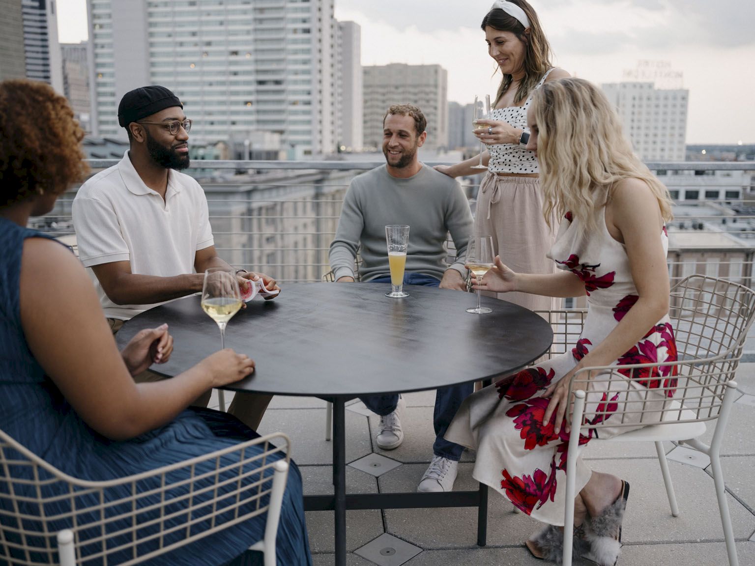 A group of five friends sits around a round rooftop table, chatting and drinking wine and juice on a sunny urban terrace.