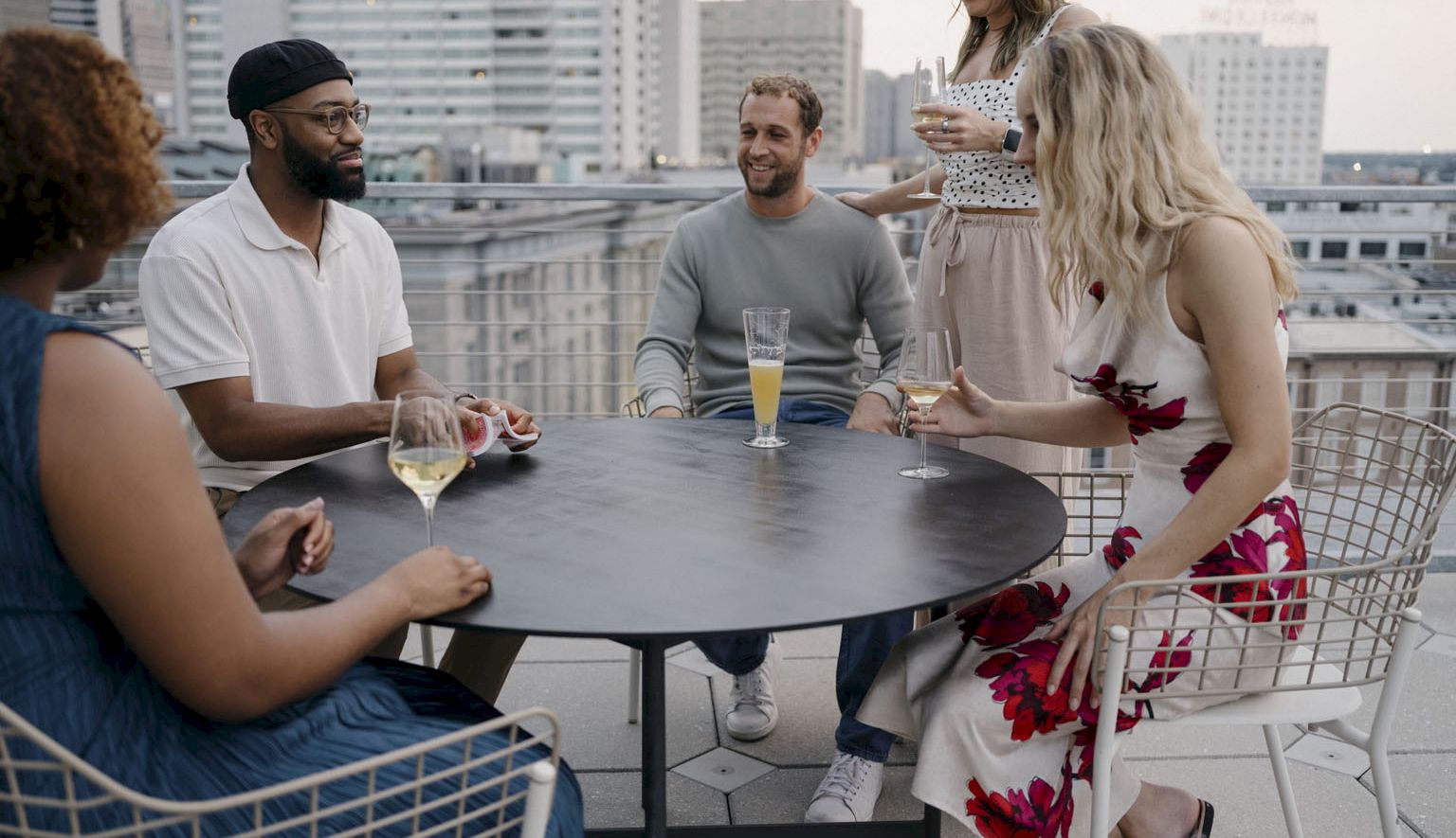 A group of five friends sits around a round rooftop table, chatting and drinking wine and juice on a sunny urban terrace.
