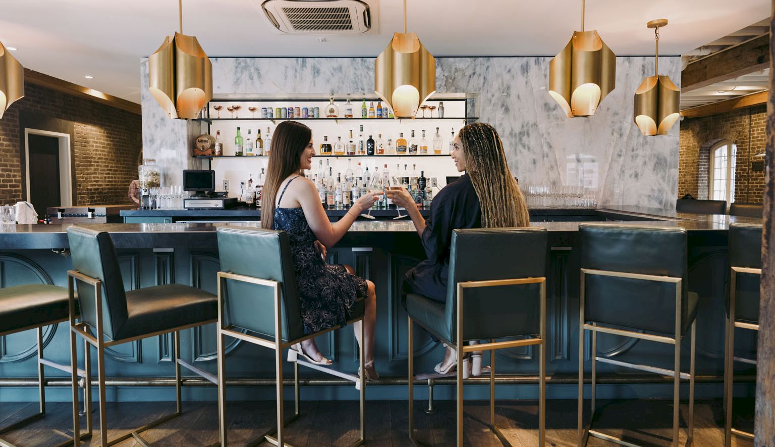 Two women sit at a modern bar chatting, bottles lined up behind, warm pendant lights, teal stools, and chic d&eacute;cor.