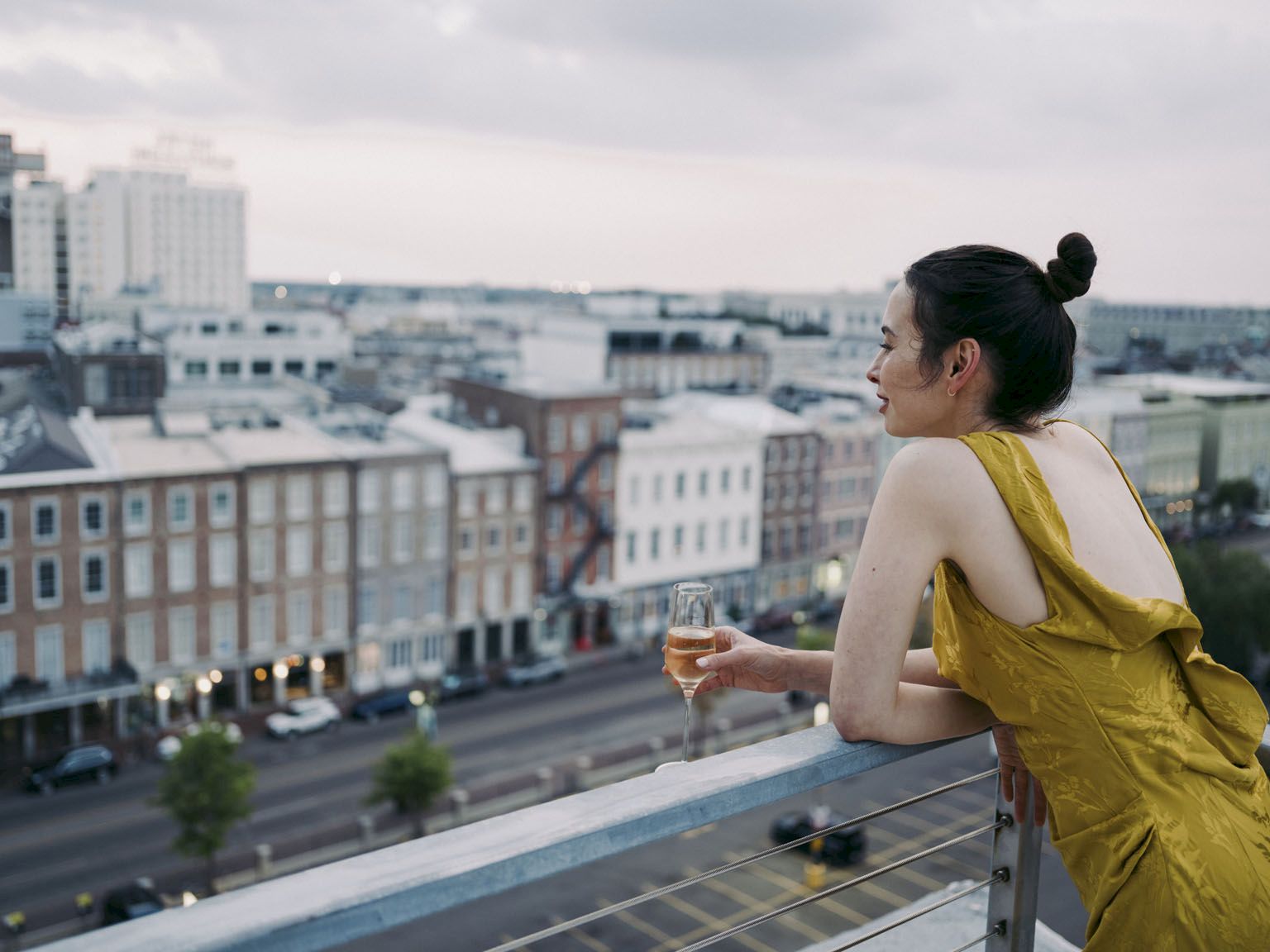 A woman in a yellow dress sips a glass of wine on a balcony, overlooking a city skyline at dusk.