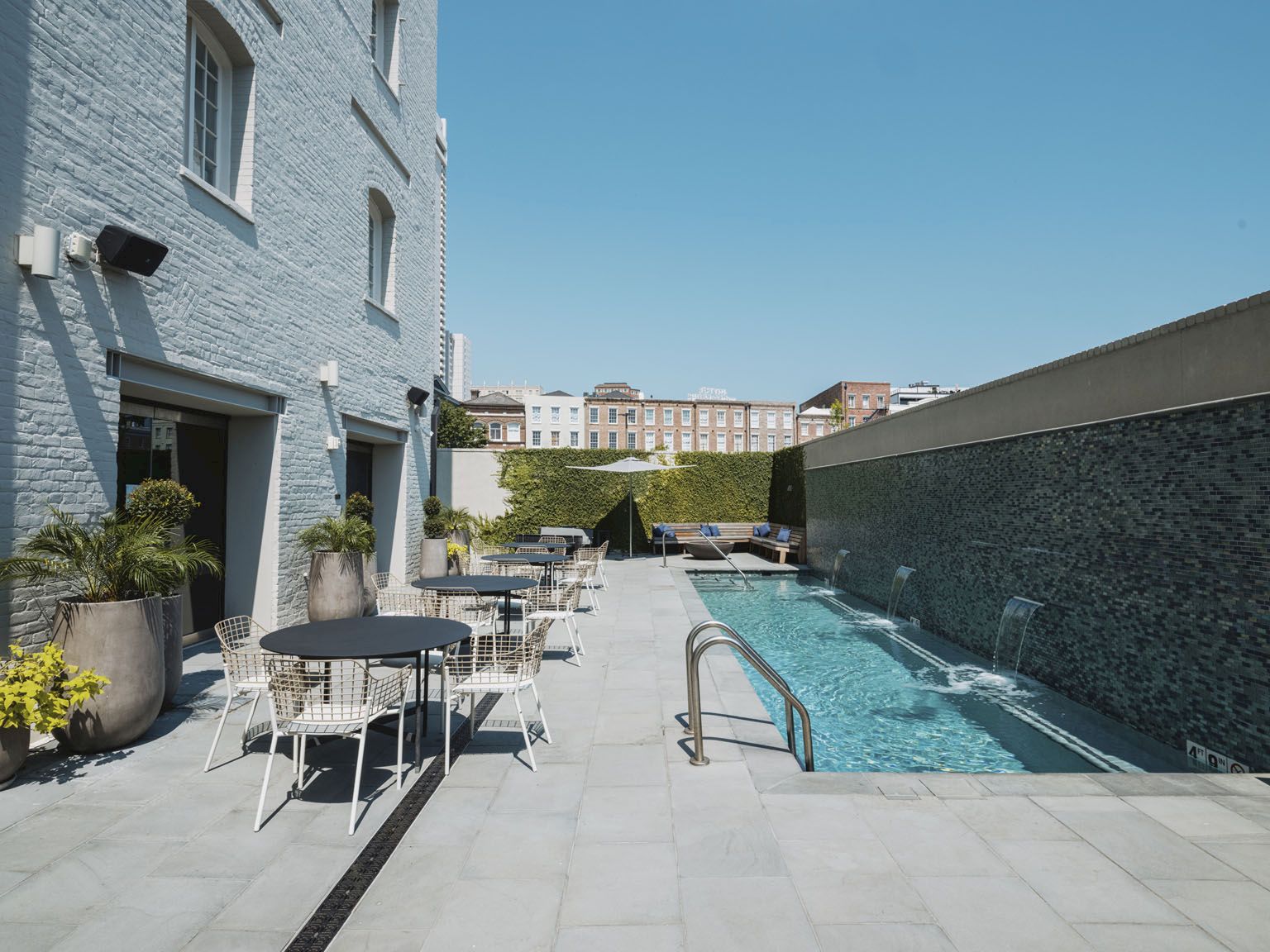 Outdoor hotel pool area with a long rectangular waterway, white courtyard furniture, potted plants, and light blue building walls under a clear sky.