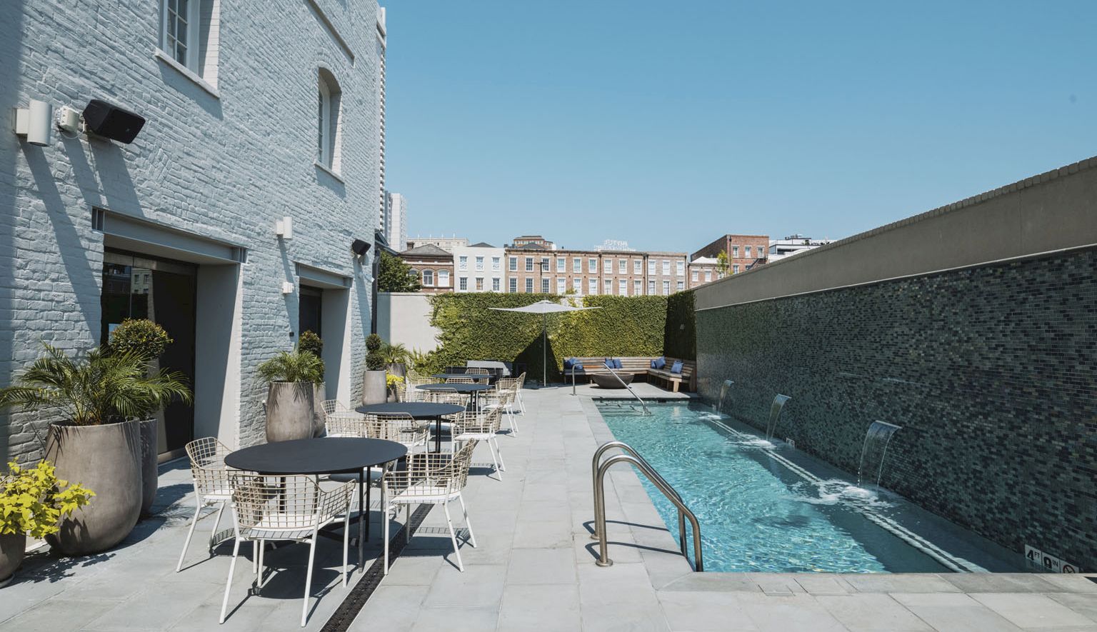 Outdoor hotel pool area with a long rectangular waterway, white courtyard furniture, potted plants, and light blue building walls under a clear sky.