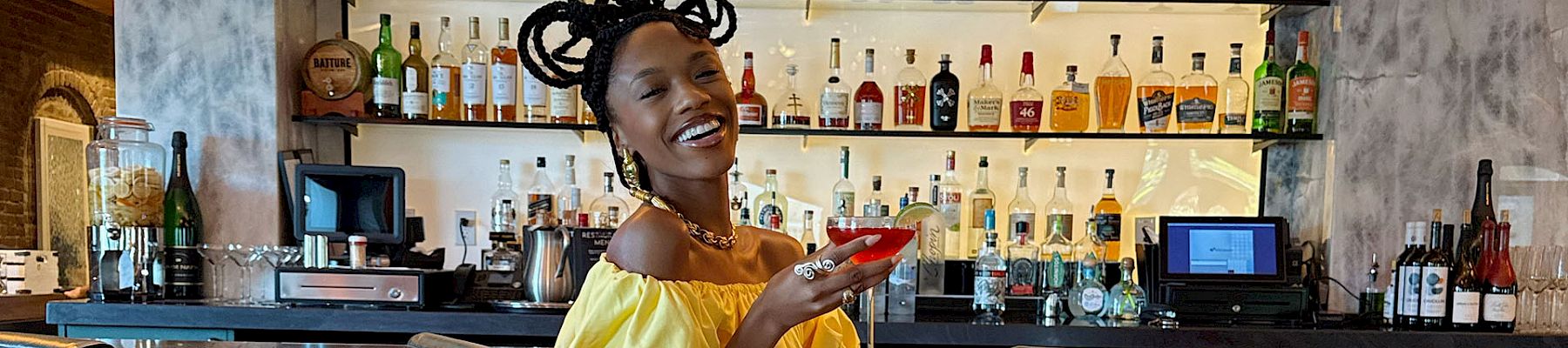 A stylish woman in a bright yellow top and pink skirt sits at a chic bar, smiling as she shows her phone near teal stools and warm lighting.