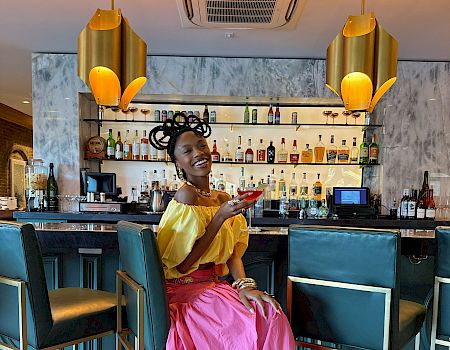 A stylish woman in a bright yellow top and pink skirt sits at a chic bar, smiling as she shows her phone near teal stools and warm lighting.