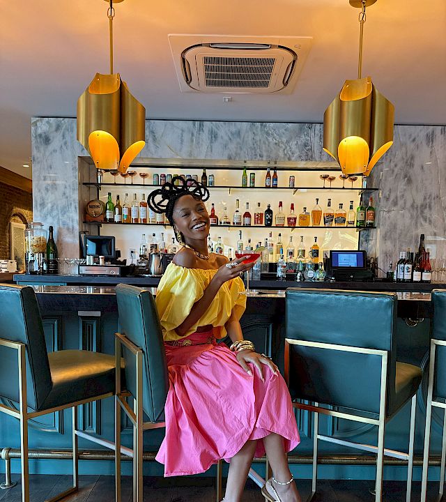 A stylish woman in a bright yellow top and pink skirt sits at a chic bar, smiling as she shows her phone near teal stools and warm lighting.