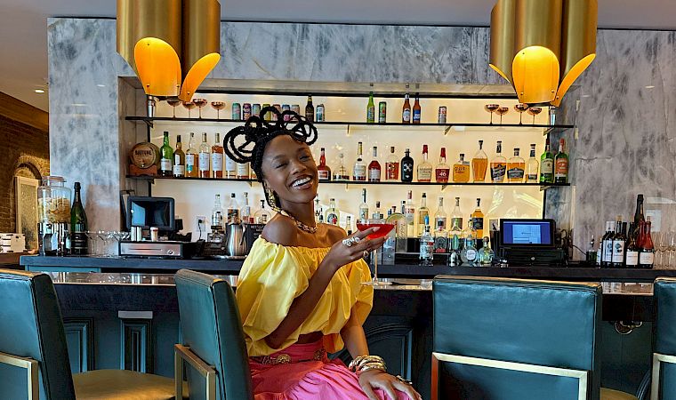 A stylish woman in a bright yellow top and pink skirt sits at a chic bar, smiling as she shows her phone near teal stools and warm lighting.