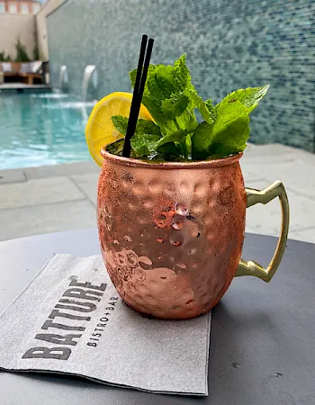 A copper mug with a minty drink garnished with lime and a straw sits on a napkin by a pool, a refreshing summer scene.