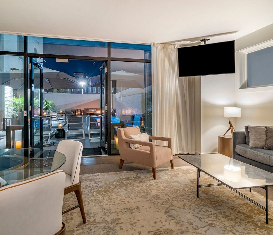 A modern hotel lounge with a sofa, coffee table, armchair, and dining table, featuring glass doors leading to an outdoor patio at night.