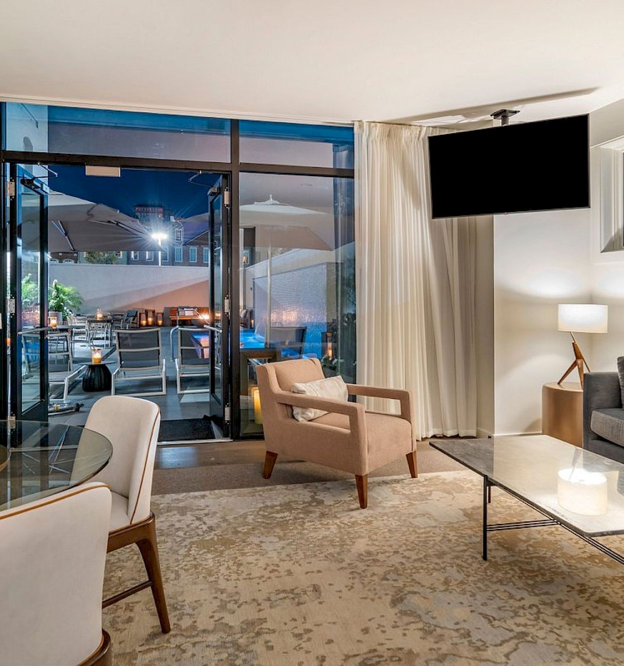A modern living room with sofas and a coffee table, opening through glass doors to an outdoor dining patio at night.