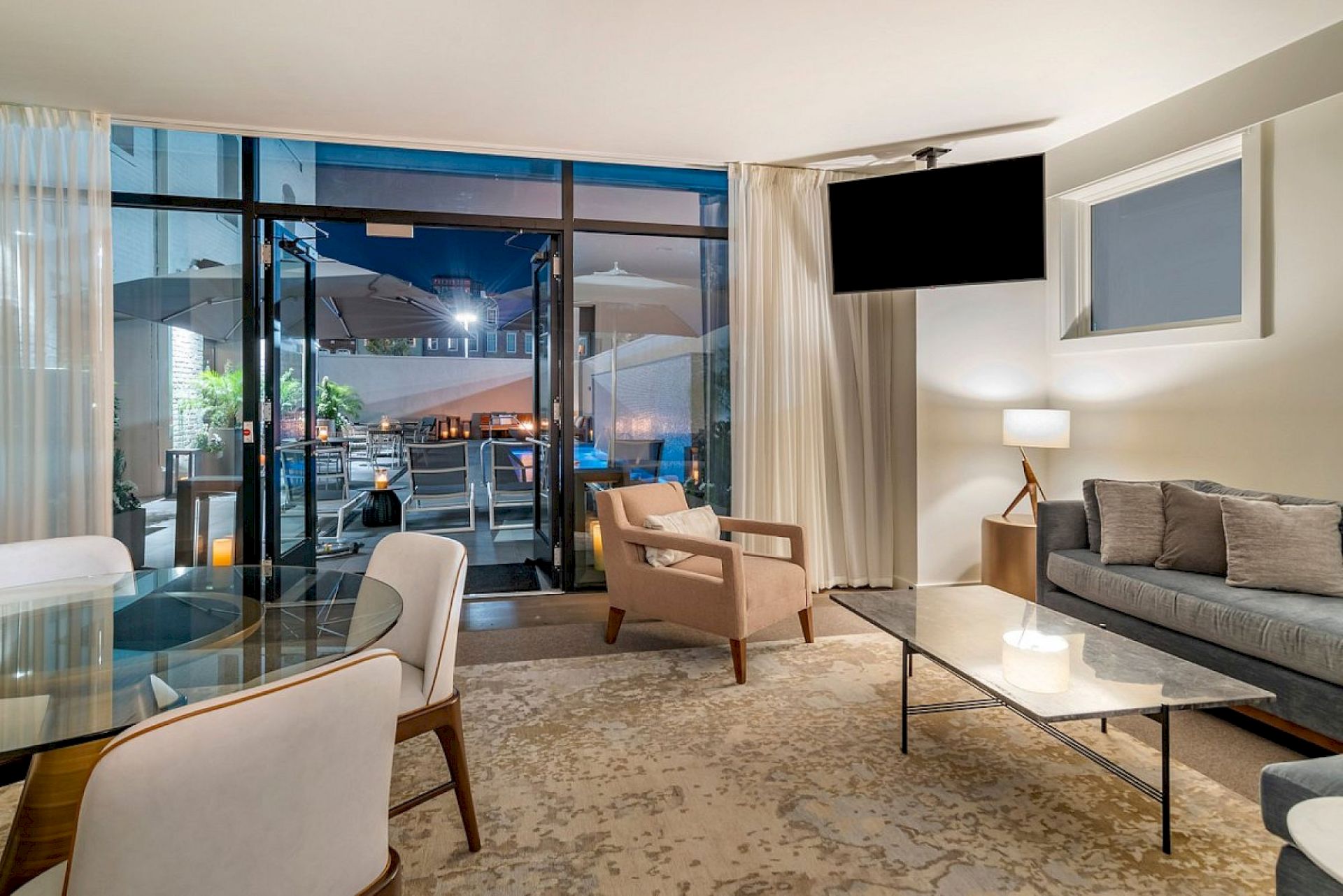 Modern living room with beige furniture, a large TV, and glass doors opening to a patio with outdoor dining and lights.