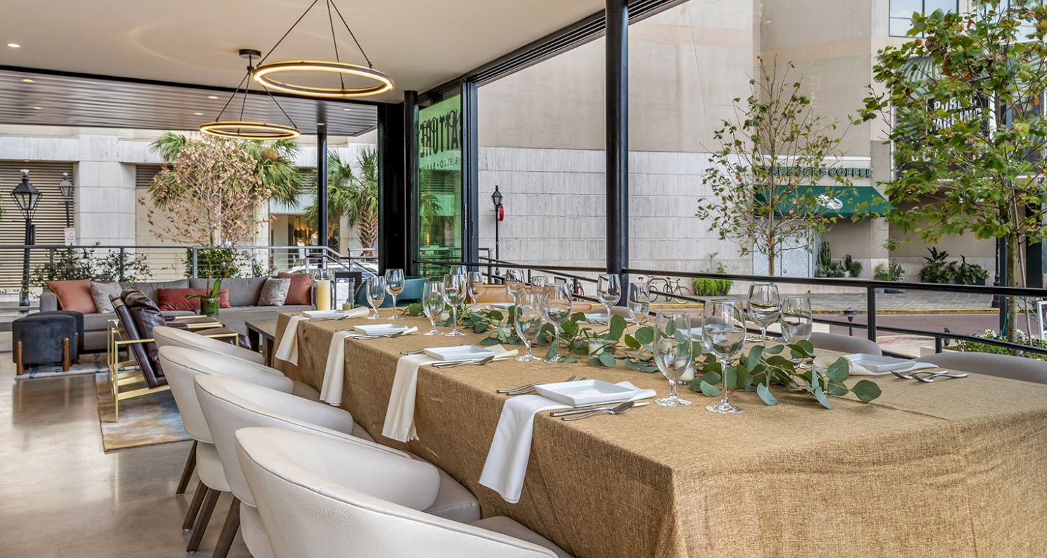 A dining area set with a long table, white chairs, and glassware, with greenery and burlap tablecloth.