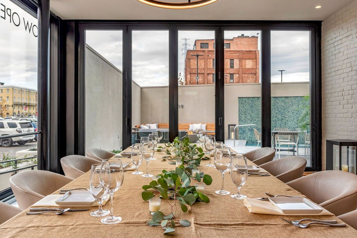 Sunlit dining room with set table, greenery centerpiece, beige chairs, and large windows/terrace showing red-brick buildings and sky.