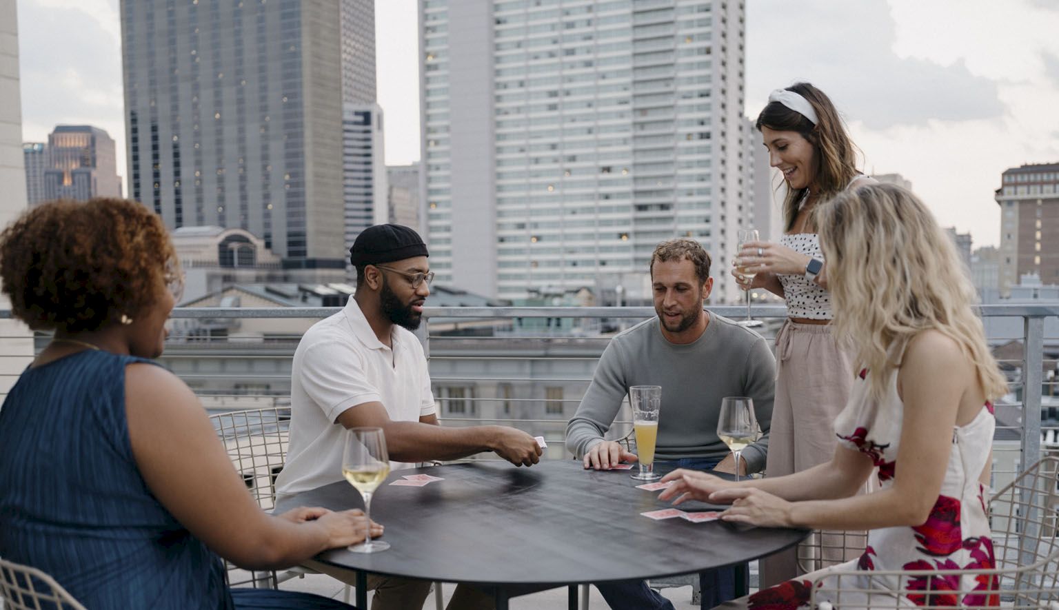 Friends play cards and sip drinks on a rooftop with skyscrapers behind them.