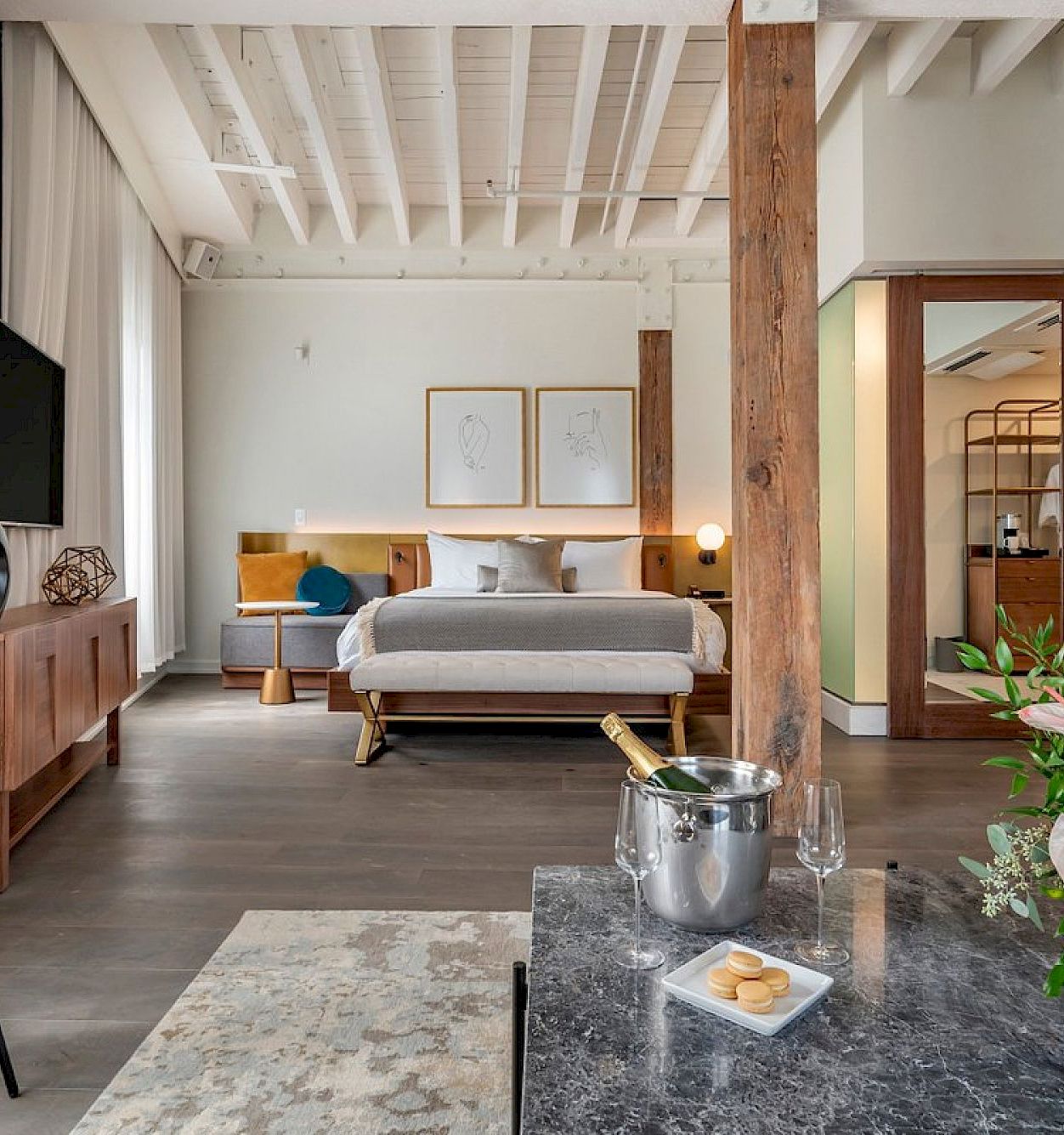 A modern hotel suite with a bed, wall art, TV, tripod floor lamp, and a vase on the marble table.