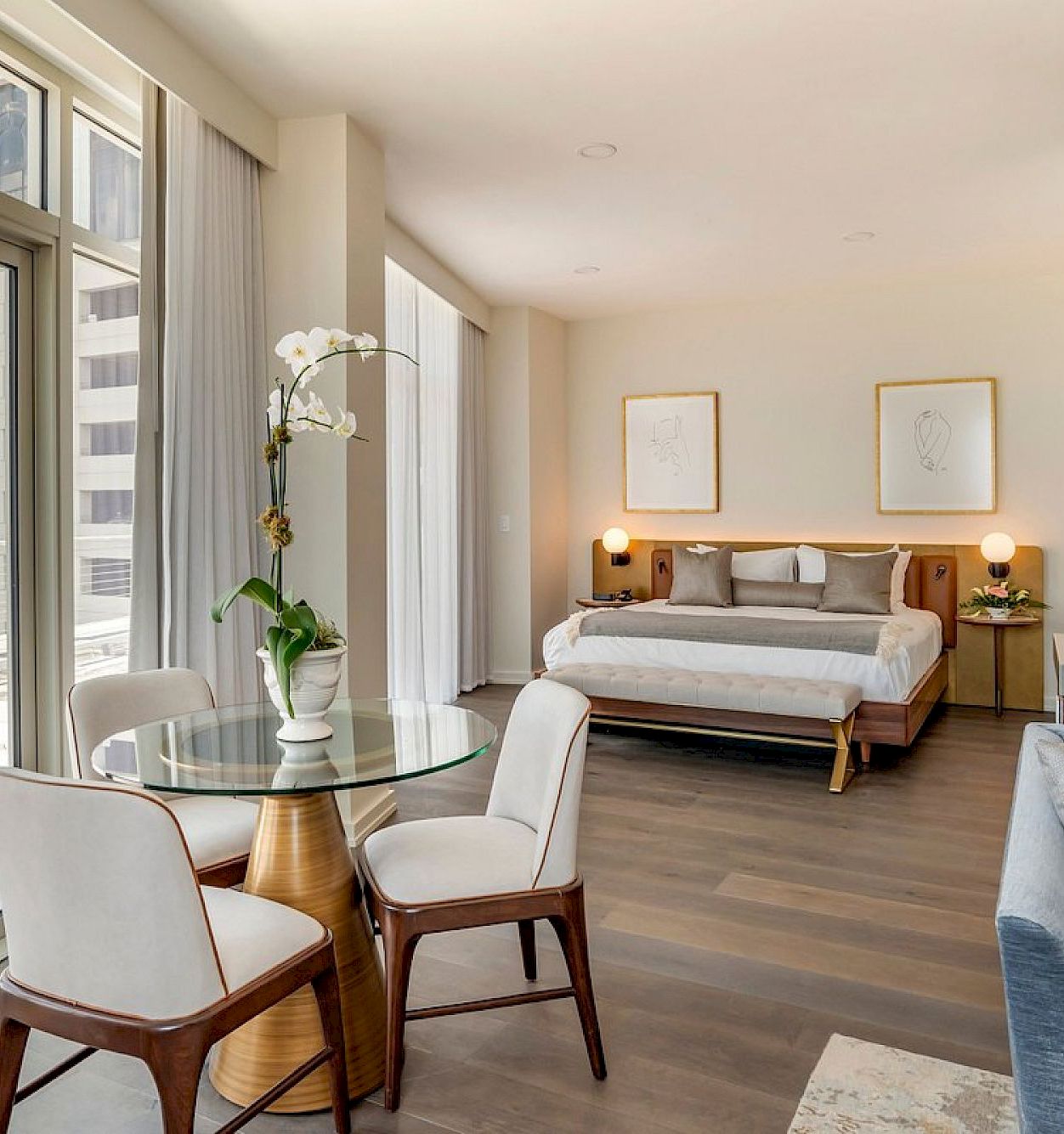 Bright modern hotel room with a bed, sofa, glass table, white curtains, and an orchid by large windows overlooking a city.
