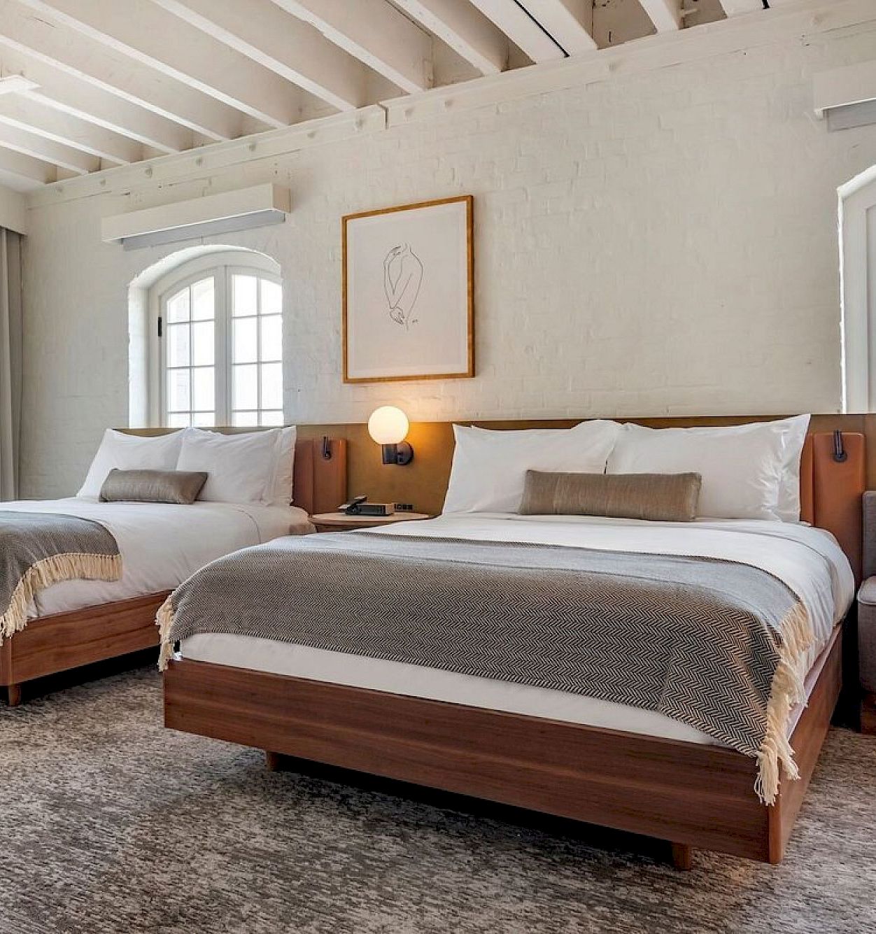 A bright modern bedroom with two twin beds, white brick walls, arched windows, warm lamps, and neutral pillows.