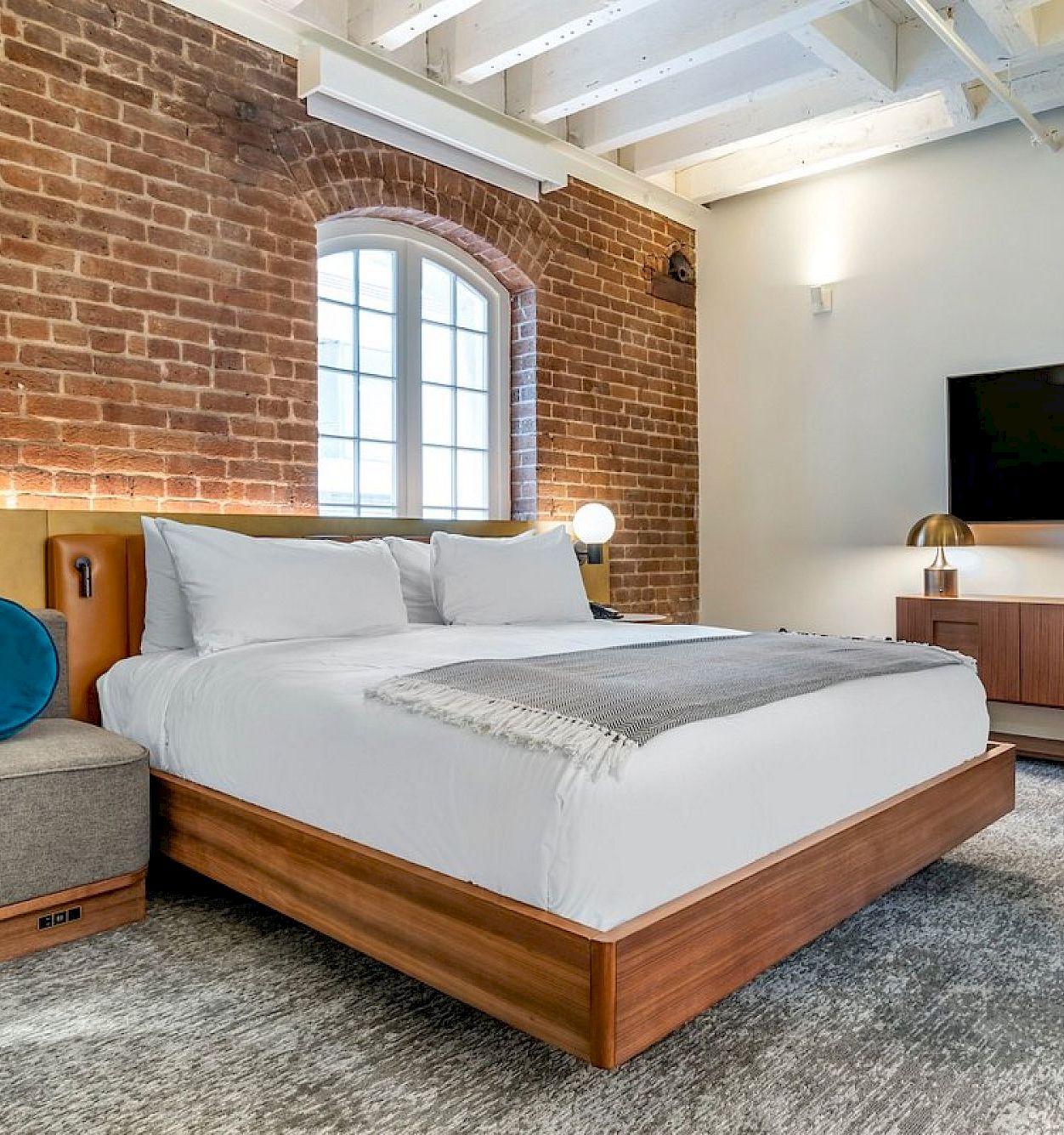 A modern loft bedroom with brick walls, a large arched window, a cozy bed, and a flat-screen TV.
